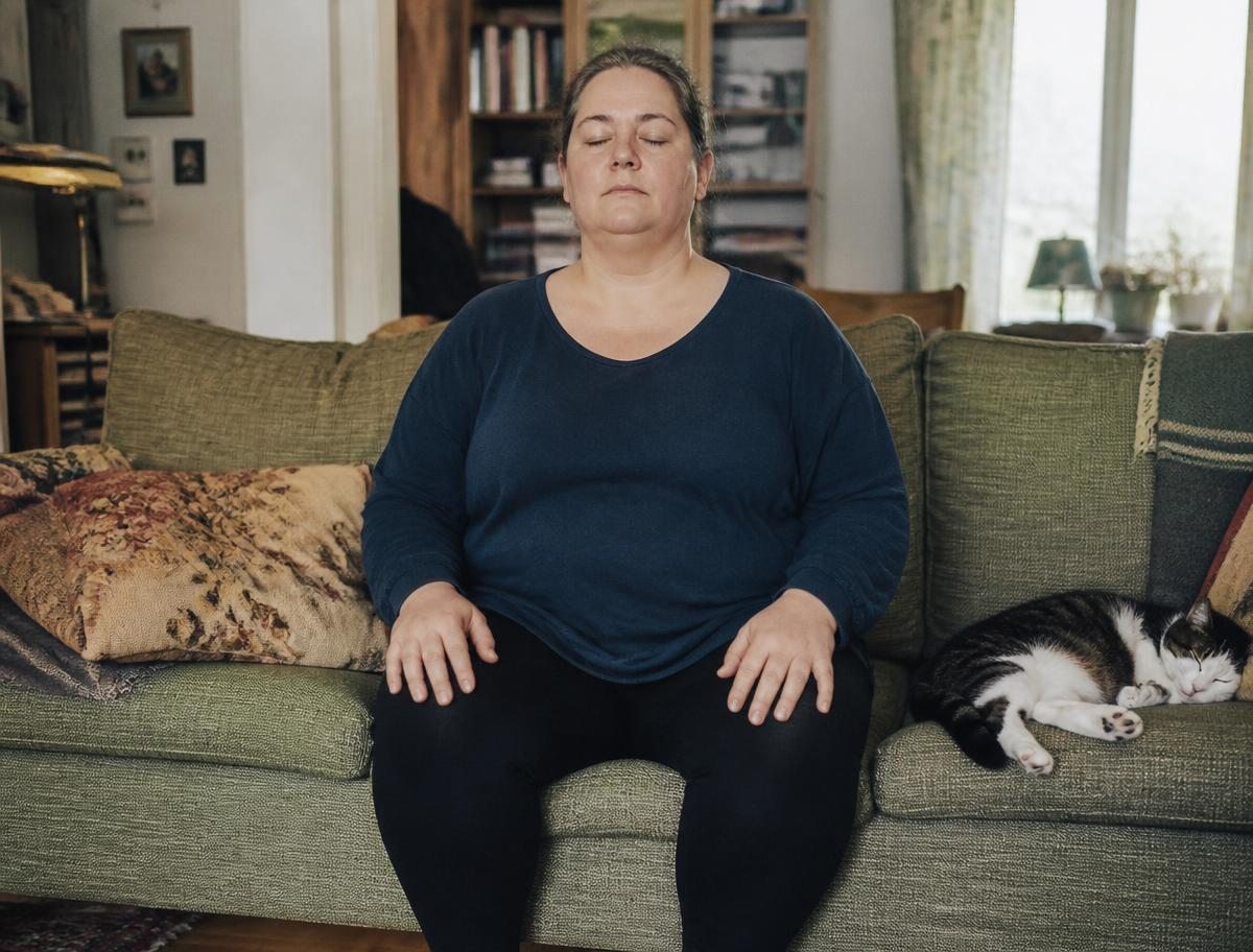 Kvinna sitter i soffan med händerna på knäna och andas lugnt, katten ligger intill